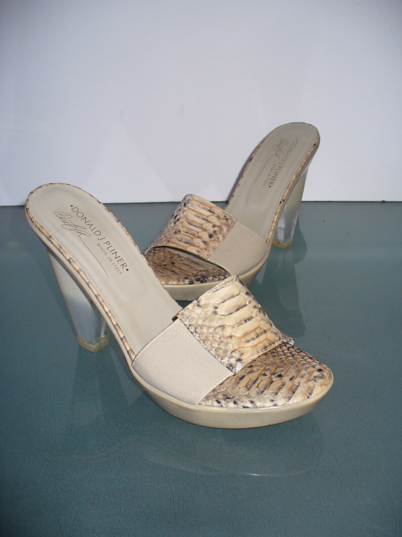 Puede incluir: Un par de mulas Donald J Pliner con punta abierta. Los zapatos presentan un estampado de piel de serpiente en la parte superior, una correa el&aacute;stica beige y tacones de acr&iacute;lico transparente. Las suelas son beige, hechas en Italia.