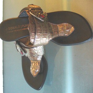 May include: A pair of thong sandals with dark brown footbeds and metallic bronze-toned straps featuring a textured crocodile-skin pattern. Each strap includes a decorative buckle with a small red stone. The text DONALD J PLINER is printed on the heel.