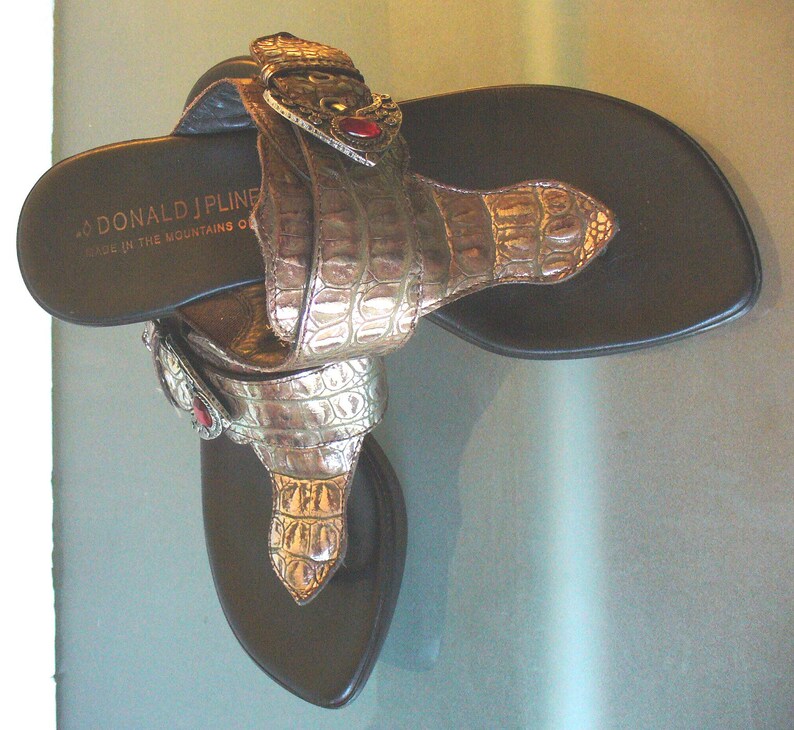May include: A pair of thong sandals with dark brown footbeds and metallic bronze-toned straps featuring a textured crocodile-skin pattern. Each strap includes a decorative buckle with a small red stone. The text DONALD J PLINER is printed on the heel.
