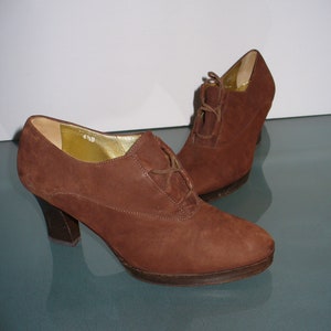 May include: A pair of brown suede lace-up shoes with a rounded toe, a stacked heel, and a platform sole. The interior lining is gold-colored, and the size 6.5 B is printed on the inner side of the shoe. The shoes feature thin brown laces.