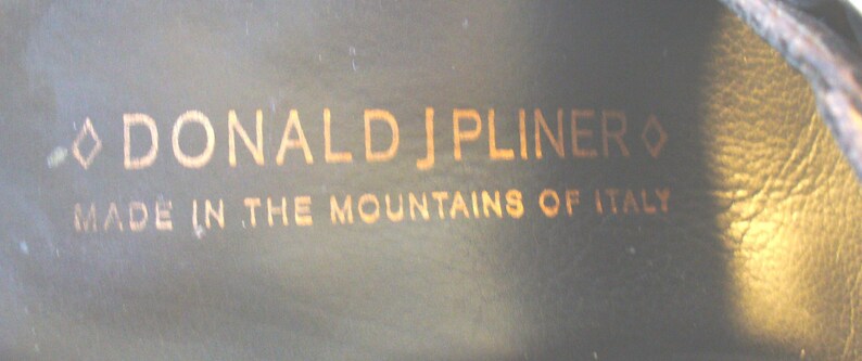May include: A dark interior surface featuring the gold-colored text DONALD J PLINER with a diamond symbol on each side, followed by the text MADE IN THE MOUNTAINS OF ITALY printed in a clean, sans-serif font on a smooth, dark material.