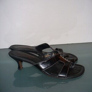 May include: A pair of black leather slide sandals with a kitten heel, featuring a textured finish on the upper straps, white contrast stitching, and a thong-style toe post. The interior footbed is marked with the Donald J Pliner brand name.