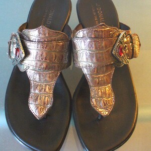 May include: A pair of bronze-colored thong sandals featuring a textured faux-alligator skin pattern on the wide strap. Each sandal includes a decorative silver-tone buckle accent with a single embedded red stone. The brand name Donald J is visible.