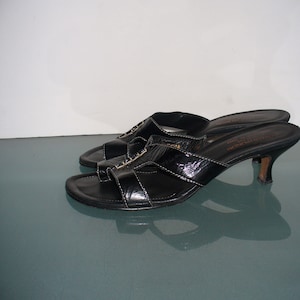 May include: A pair of black patent leather slide sandals with a low kitten heel, an open toe, and a thong-style strap across the front. The footbed features the text Donald Pliner and Made in Italy printed in gold lettering on the heel area.
