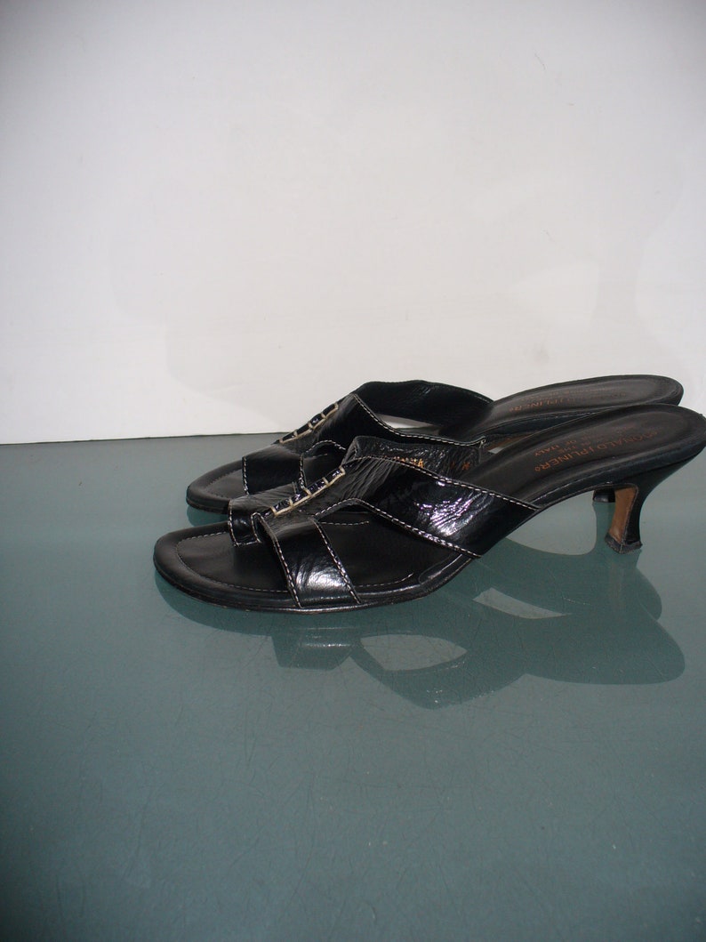 May include: A pair of black patent leather slide sandals with a low kitten heel, an open toe, and a thong-style strap across the front. The footbed features the text Donald Pliner and Made in Italy printed in gold lettering on the heel area.