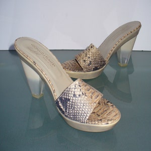 Puede incluir: Un par de sandalias deslizantes de punta abierta Donald J Pliner. Las sandalias presentan un estampado de piel de serpiente en tonos beige, marr&oacute;n y negro. Tienen tacones transparentes y gruesos y una suela de plataforma. El nombre de la marca es visible en la plantilla.