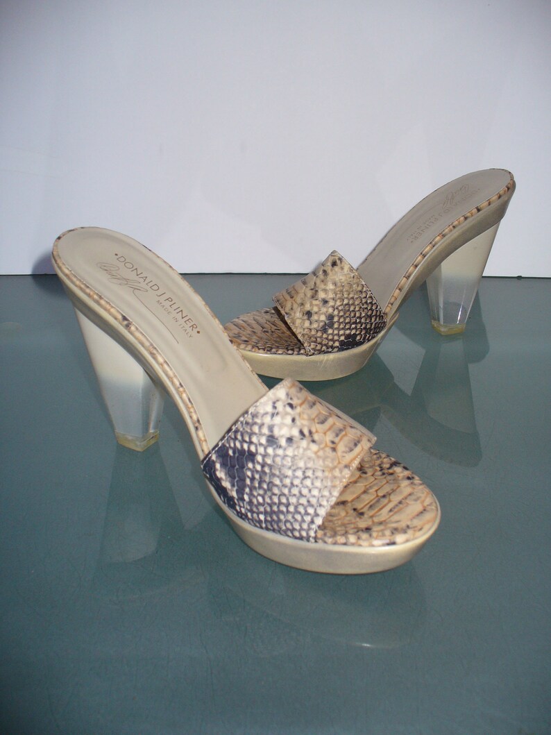 Puede incluir: Un par de sandalias deslizantes de punta abierta Donald J Pliner. Las sandalias presentan un estampado de piel de serpiente en tonos beige, marr&oacute;n y negro. Tienen tacones transparentes y gruesos y una suela de plataforma. El nombre de la marca es visible en la plantilla.