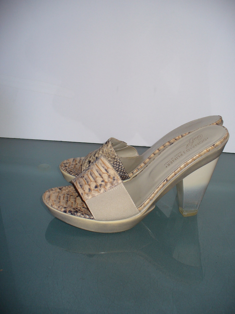 Puede incluir: Un par de sandalias de tac&oacute;n alto con punta abierta. Las sandalias presentan un estampado de piel de serpiente en tonos beige, marr&oacute;n y negro. Los tacones son transparentes y angulares, y las suelas son beige. La marca "DONALD J. PLINER MADE IN ITALY" es visible.