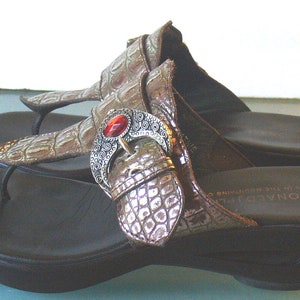 May include: A pair of brown thong sandals with a faux crocodile skin texture on the straps. Each sandal features a decorative silver-toned metal buckle ornament centered with an oval red stone. The sandals have a black footbed and a wedge heel.