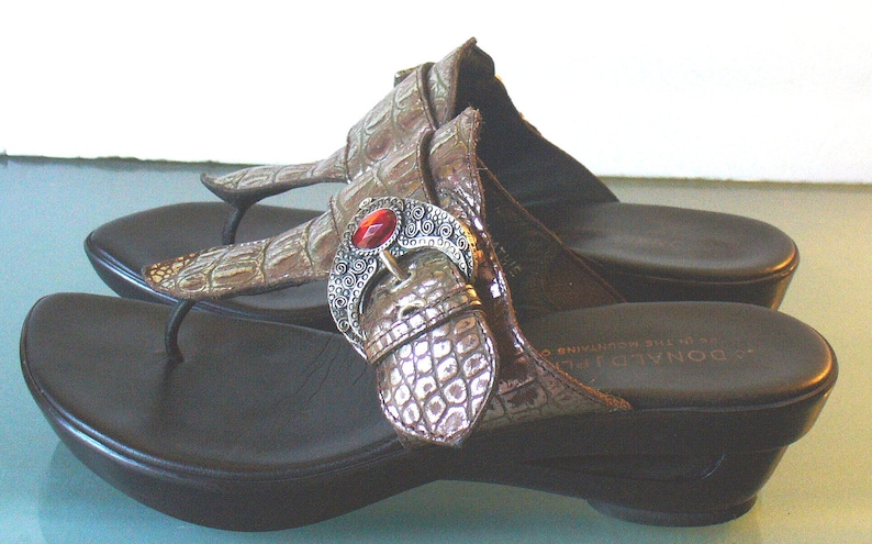May include: A pair of brown thong sandals with a faux crocodile skin texture on the straps. Each sandal features a decorative silver-toned metal buckle ornament centered with an oval red stone. The sandals have a black footbed and a wedge heel.