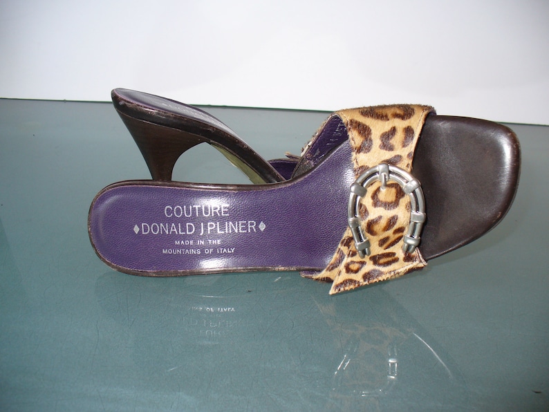 Puede incluir: Sandalia abierta marr&oacute;n y con estampado de leopardo con hebilla plateada. La sandalia tiene una plantilla morada con el texto "COUTURE DONALD J PLINER MADE IN THE MOUNTAINS OF ITALY". El tac&oacute;n es marr&oacute;n oscuro.