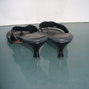 May include: A pair of black leather thong sandals featuring a low kitten heel, a wide strap across the top of the foot, and a smooth insole. The sandals have a dark finish and a curved heel design, showing the back and side profile of the footwear.