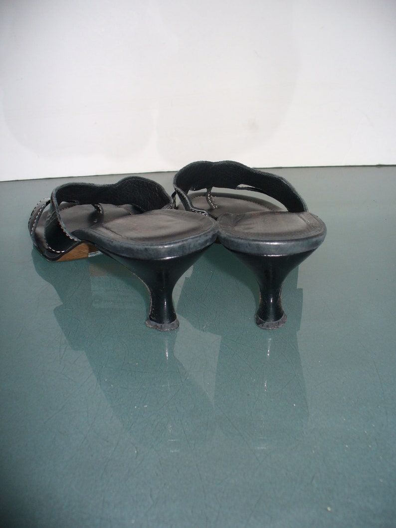May include: A pair of black leather thong sandals featuring a low kitten heel, a wide strap across the top of the foot, and a smooth insole. The sandals have a dark finish and a curved heel design, showing the back and side profile of the footwear.