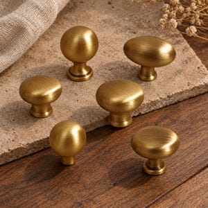 Puede incluir: Un conjunto de seis pomos para armarios de metal en tono latón con acabado cepillado. El set incluye cuatro pomos redondos y dos pomos ovalados, cada uno con una base circular y una superficie superior lisa y redondeada para muebles.