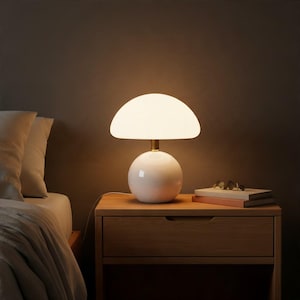 Peut inclure: Une lampe de table en forme de champignon avec une base sphérique blanche lisse et un large abat-jour en dôme de verre blanc lumineux. La lampe présente un col fin en métal doré reliant la base à l'abat-jour, diffusant une lumière douce.