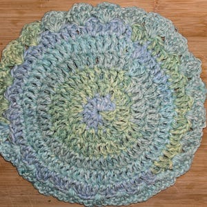 May include: A round, hand-crocheted coaster or doily featuring a variegated yarn pattern in soft shades of light blue, mint green, and white. The circular textile has a textured, spiraling stitch design with a scalloped, decorative edge.