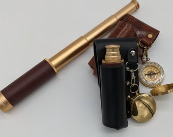 Könnte beinhalten: Ein goldfarbenes Teleskop mit einer braunen Ledertasche, einem Kompass und einer kleinen Flasche mit einer schwarzen Ledertasche. Das Teleskop ist scharf gestellt, die anderen Gegenstände sind leicht unscharf.