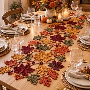 May include: A crochet table runner featuring a repeating pattern of maple leaves in shades of burgundy, orange, yellow, and olive green, laid across a wooden dining table set with white plates, linen napkins, and clear glassware.