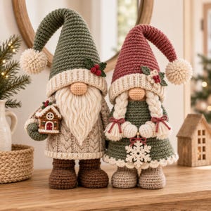 May include: Two crochet gnomes with cream-colored pom-poms on their hats. The left gnome wears a green hat and beige sweater, holding a small house. The right gnome wears a red hat and green dress with white braided hair and a snowflake ornament.