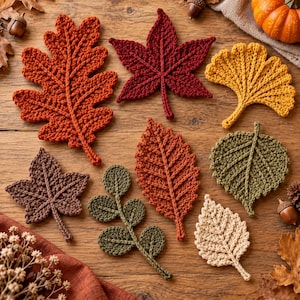 May include: A collection of eight crocheted leaves in various shapes and autumn colors, including orange, deep red, yellow, brown, olive green, and cream, arranged on a wooden surface with small acorns and a pumpkin.