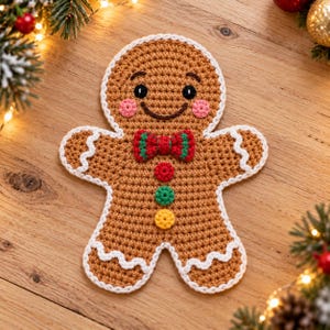 May include: A tan crochet gingerbread man ornament featuring a white scalloped trim, a red and green bowtie, and three small buttons in red, green, and yellow. The figure has black eyes, a brown stitched smile, and pink circular cheeks.