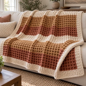 May include: A waffle-knit throw blanket featuring horizontal stripes in cream, tan, and rust orange. The blanket has a thick, solid cream-colored border along all edges, showcasing a textured grid pattern throughout the entire fabric.