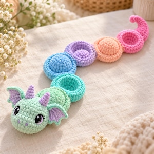 May include: A crocheted dragon toy with a mint green head, purple horns, and small wings. The body is composed of a series of connected, circular, plush segments in alternating colors of mint green, teal, blue, purple, peach, and pink.