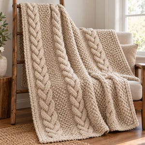 May include: A beige knit throw blanket featuring thick vertical cable knit patterns alternating with textured seed stitch sections. The blanket is draped over a wooden ladder and a chair, showcasing a chunky yarn construction.