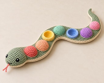 May include: A crocheted snake toy in sage green with a cream-colored underside. The snake features a series of six circular, bowl-shaped indentations along its back in red, orange, yellow, teal, blue, and purple. It has black eyes and a red tongue.