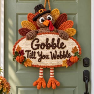 May include: A crocheted turkey wall hanging featuring a brown turkey wearing a black pilgrim hat with a yellow buckle. The turkey holds an oval cream-colored sign with the brown text Gobble Till You Wobble, decorated with small orange pumpkins and autumn leaves.