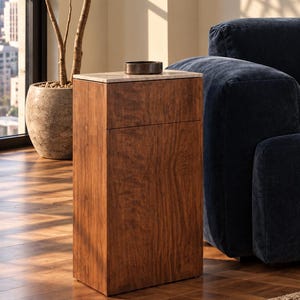 May include: A tall, rectangular wooden side table with a rich brown wood grain finish and a flat, light-colored stone top. A small, dark, circular bowl sits centered on the stone surface of the table, which features a clean, vertical design.