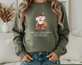 May include: A military green crewneck sweatshirt featuring a graphic of a disheveled Santa Claus holding a sack. The text reads: Budget cuts sold the reindeer, carrying the sack Merry Christmas! The garment has long sleeves and a ribbed neckline.