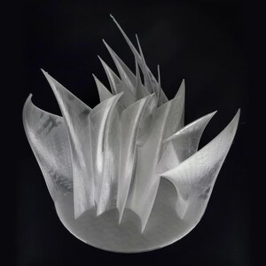 May include: A translucent, white, 3D-printed sculpture featuring a circular base with multiple sharp, upward-pointing, blade-like fins that curve and intersect to create a complex, layered, and abstract geometric structure.