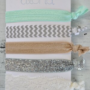 May include: A set of five hair ties on a white card with the word "Aloha" printed on it. The hair ties are in a variety of colors and patterns, including mint green, gray and white chevron, beige, silver glitter, and white lace.