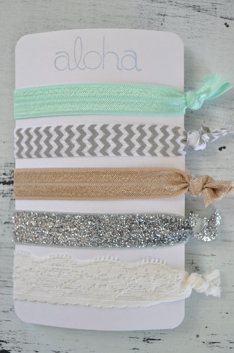 May include: A set of five hair ties on a white card with the word "Aloha" printed on it. The hair ties are in a variety of colors and patterns, including mint green, gray and white chevron, beige, silver glitter, and white lace.
