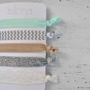 May include: A set of five hair ties on a white card with the word "Aloha" printed on it. The hair ties are in a variety of colors and patterns, including mint green, gray and white chevron, beige, silver glitter, and white lace.