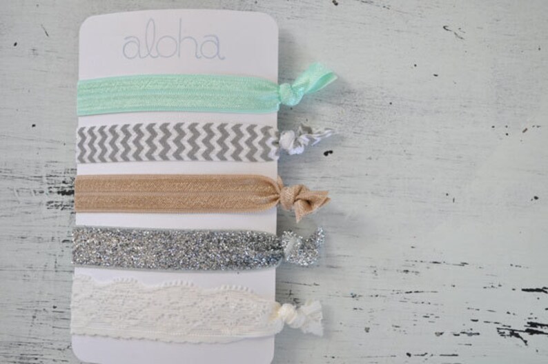 May include: A set of five hair ties on a white card with the word "Aloha" printed on it. The hair ties are in a variety of colors and patterns, including mint green, gray and white chevron, beige, silver glitter, and white lace.