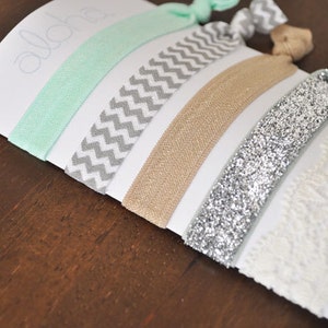 May include: A set of five hair ties in various colors and patterns, including mint green, gray chevron, beige, silver glitter, and white lace. The hair ties are displayed on a white card with the word "Aloha" written on it.