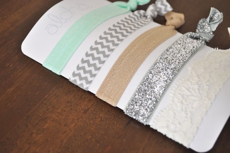 May include: A set of five hair ties in various colors and patterns, including mint green, gray chevron, beige, silver glitter, and white lace. The hair ties are displayed on a white card with the word "Aloha" written on it.