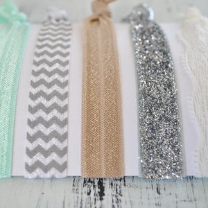 May include: Set of five hair ties in various colors and patterns. The hair ties include a mint green, a gray and white chevron, a beige, a silver glitter, and a white lace.