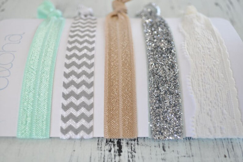 May include: Set of five hair ties in various colors and patterns. The hair ties include a mint green, a gray and white chevron, a beige, a silver glitter, and a white lace.