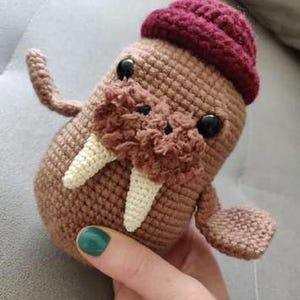 Puede incluir: Un pequeño peluche de morsa tejido a ganchillo en color marrón, con un gorro granate, dos colmillos blancos, ojos de plástico negro y un hocico texturizado. El juguete tiene dos pequeñas aletas que sobresalen a los lados.