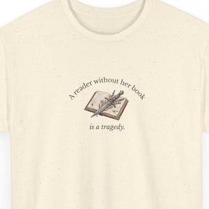Może przedstawiać: Kremowy t-shirt z krótkim rękawem o nakrapianej fakturze. Na środku znajduje się ilustracja otwartej książki z leżącym na niej mieczem, otoczona tekstem: A reader without her book is a tragedy.