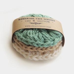 May include: Handmade face scrubby made from 100% cotton. The scrubby is a light green, beige, and white crocheted round pad. The scrubby is wrapped in brown paper with the text "Handmade Face Scrubby - 100% Cotton - www.rightsoap.com".