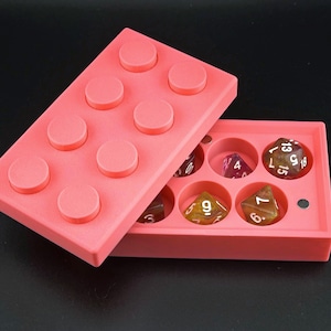 May include: A coral-colored dice box, designed to resemble a building block, with a lid and a compartment holding several multi-sided dice. The box is open, revealing the dice in various colors and shapes. The background is black.