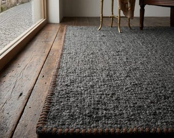 May include: A rectangular area rug with a thick, dark grey, textured weave and a contrasting brown stitched border. The rug is positioned on a dark wooden floor, extending from the foreground towards the back of the room.