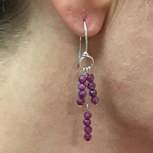 May include: A silver earring featuring a hook wire and a silver ring from which three short strands of small, faceted, deep pinkish-purple gemstone beads hang. The beads are arranged in two shorter side strands and one longer center strand.