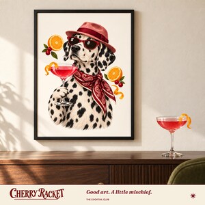 May include: A framed wall art print featuring a Dalmatian dog wearing a red fedora, red sunglasses, and a red paisley bandana. The dog holds a pink cocktail glass, surrounded by orange slices and cherries. The print is displayed above a wooden surface.