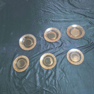 May include: A set of six round glass saucers featuring a dark amber center and a wide, metallic gold rim decorated with an intricate, embossed floral and geometric pattern along the outer edge of each individual piece.