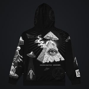 May include: A black hoodie featuring a graphic print of a cracked pyramid with an eye, smoke, and UFOs. The design includes a person standing in a light beam, celestial elements, and the text "COORDINATES UNKNOWN." A QR code is visible on the sleeve.